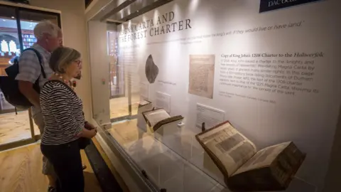 A man and a woman look into a glass case in a museum housing two old books. A sign behind the display reads: "MAGNA CARTA AND THE FOREST CHARTER".