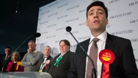 Andy Burnham, wearing a black suit, white shirt and burgundy tie with a red rosette on his lapel, speaks into a microphone next to a number of smartly-dressed fellow candidates for Greater Manchester mayor, in front of a wall with signs on it reading 'GM Elects'