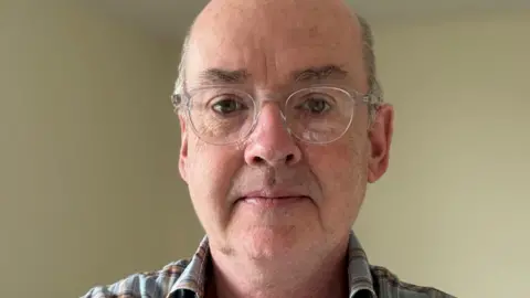 Crispin Sinclair Crispin Sinclair is looking at the camera and is balding. He is wearing spectacles and a checked shirt