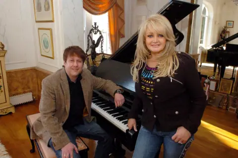 Rob Marshall A man with brown hair, wearing blue jeans, a black t shirt and a brown jacket. He is sat at a piano. Stood up beside him is a glamorous blonde woman wearing blue jeans and a black top and jacket. They are both looking at the camera and smiling.