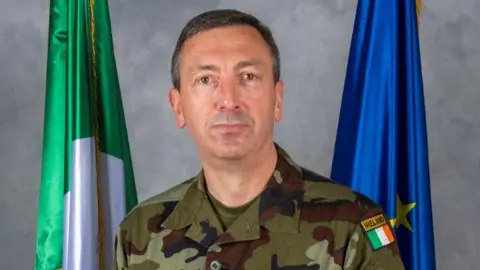 Irish Defence Forces A man in military uniform with the Irish flag on his shoulder. He is looking at the camera with a neutral expression. Behind him are the flags of Ireland and the EU.