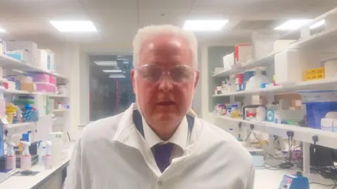 A man with white hair wearing a white lab coat and clear goggles
