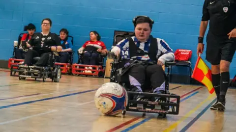 Powerchair Football champion Matt Gilbert in action. He is wearing his Albion kit of white and blue and there is a football in front of his chair. We can see the bottom half of a referee to the right of the screen. He is holding a flag. There are some other players behind Mr Gilbert. 
