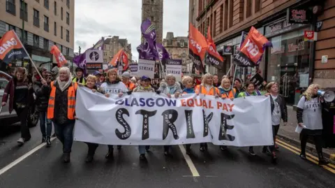 A group of workers protesting as they walk down a street, holding several banners and signs - including one saying GLASGOW EQUAL PAY STRIKE.