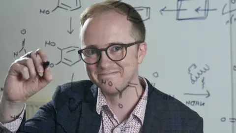 La Universidad de Edimburgo sonriendo, el profesor Stephen Wallace escribe fórmulas químicas en una mesa de visualización transparente.