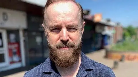 BBC A man with a beard and moustache standing on a street infront of shops. He is wearing a blue collared shirt and looking at the camera