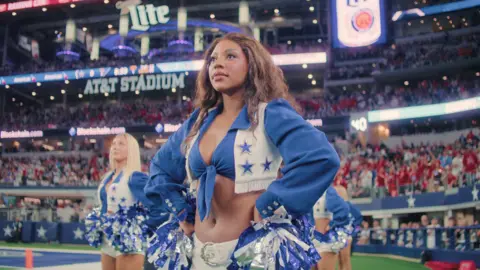 Netflix Two dancers on the field look straight ahead. The dancer in the front has long brown hair and the dancer in the back has long straight blonde hair. They wear the signature blue and white Dallas Cowboys Cheerleaders uniform 