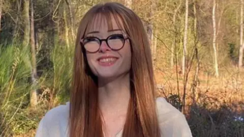 Family handout Brianna Ghey, who has long straight brown hair and wears black glasses, smiles at the camera while standing against a backdrop of woodland