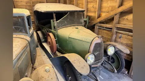 Humbert & Ellis 1924 Françon Voiturette sold for £14,000. This is a dust covered car with green paintwork and a black soft top. It has black wheels and a rusty looking hub cap attached to its bonnet. It is in the corner of a barn, against wooden walls. There is a glimpse of another vintage car on the left.