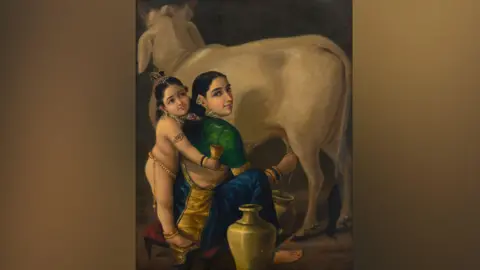 A woman wearing a green blouse and a blue saree is seen milking a cow in the oil portrait. A child, who is Hindu deity Krishna, is seen standing next to her wearing jewellery. 
