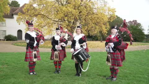 Four band members stood outside playing in their uniforms, consisting of a black velvet jacket, red tartan bagpipes, black drum, white lace collars and black hats. A tree is visible in the background