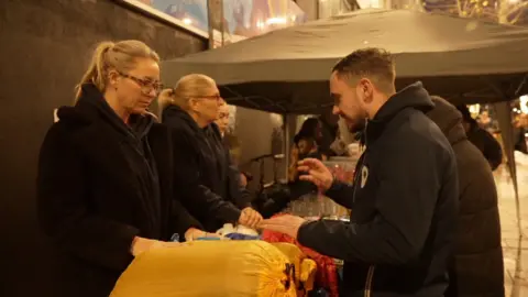 Three individuals engage in a discussion at an outdoor stall, surrounded by food and clothing items.