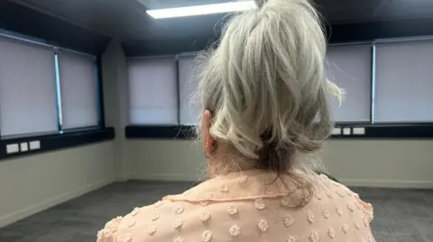 A woman is sitting in a chair with her back to the camera. Her hair is ash blonde and tied up and she's wearing a pink jacket. She's in a bare room with the blinds drawn