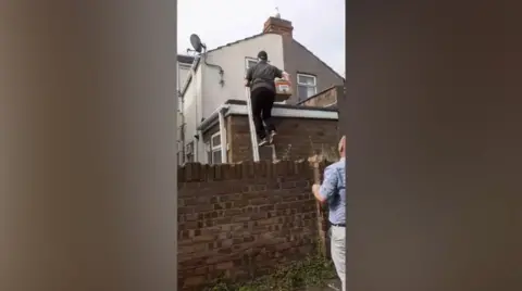 Facebook A man looks on from ground level as another man climbs a ladder lent against a flat roof. He is carrying a box.