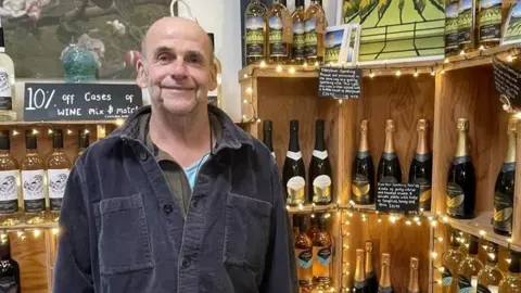 Owner John Coulson stood in the shop of the vineyard in front of wooden shelves filled with different bottles of wine. he is bald and smiling at the camera wearing a dark grey large fabric shirt. 