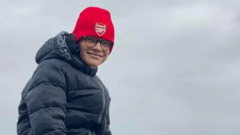 Harry Basham is sitting on a rock on a beach. He is wearing a red hat and a black hooded jacket. He is wearing black tracksuit bottoms and black and red trainers. The picture was taken on a cloudy day.