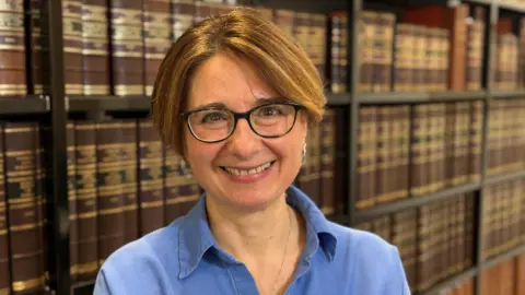 Director of Research at Cambridge University Press and Assessment, Evelina Galaczi, stands in front of a bookcase. She is smiling and wearing a blue shirt and glasses. 