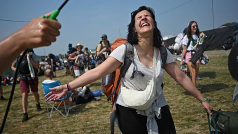 Woman in shorts and a T-shirt smiling as she is sprayed with water on a sunny day while taking bags into Glastonbury