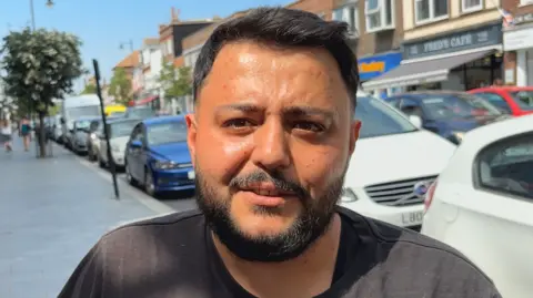 Jodie Halford/BBC Memmzy Ozy has short black hair and a beard. He is wearing a black T-shirt in front of a row of parked cars.
