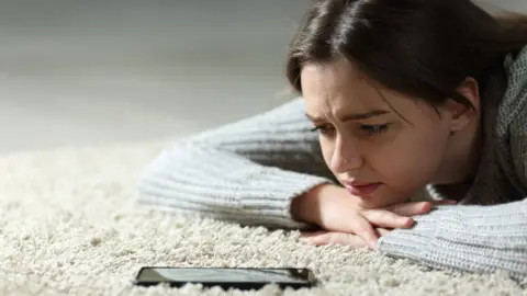 Getty Images A young woman lying on the floor looking at her smartphone with concern