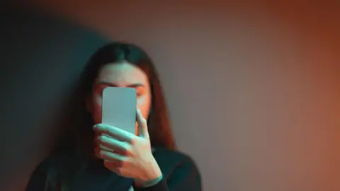 Getty Images Teenager holding a mobile phone up in front of her face. Girl has long hair and is wearing a dark-coloured jumper. Her face is not clearly visible.