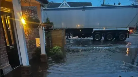 Floodwater at around ankle-level can be seen reaching the black front door of John Frosdick's home. Behind the house, on the road, a lorry can be seen making its way through the floodwaters. 
