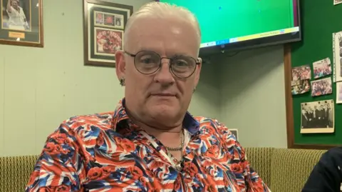 A man in a Union Jack shirt relaxes in a pub environment and smiles for the camera