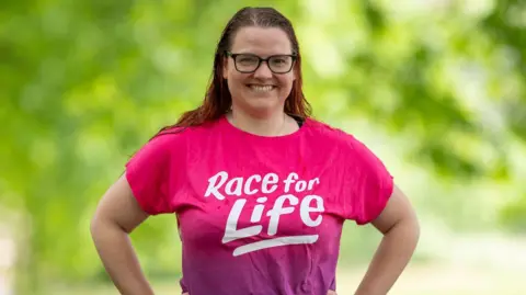 A woman wearing a pink top with "Race for Life" written on it in a white font. She is smiling and has long brown hair and black rimmed glasses.