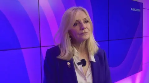 A woman wearing a purple blazer with mid length blonde hair stands in front of several tv screens