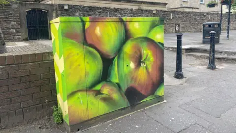 A Junction box on a street. There is a brick wall right beside it and behind it there is other street furniture like bollards and a bin. The junction box has been painted with large green apples.