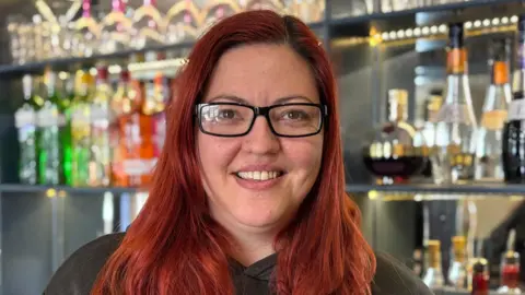 Jodie Halford/BBC Hayley Mann has red hair and glasses. She is smiling behind the bar of her pub, which has bottles of drink and glasses stacked on a shelf.