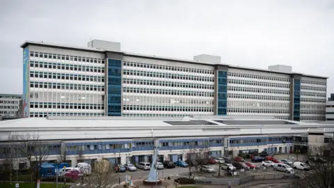 Getty Images The outside of a large hospital, which is tall, blue and white with many windows, and car parked in front of it.