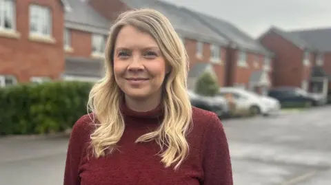 Kayleigh Roper Kayleigh Roper is standing on a residential street with red brick houses in the background. The background is blurred but she is in focus. There are a few cars and a hedge behind her. She has long wavy blond hair in a side parting and is smiling into camera. She is wearing a dark red polo neck dress.