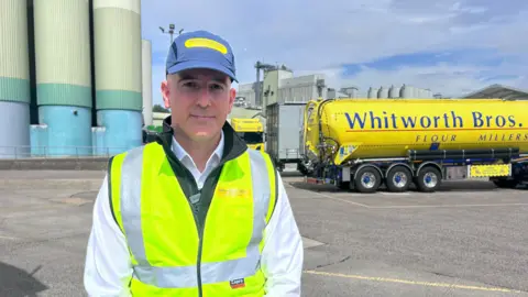 Kate Bradbrook/BBC Neil Fyfe, standing outside, wearing a cap, white jacket, high-vis jacket, looking at the camera, with a lorry behind him to the right. 