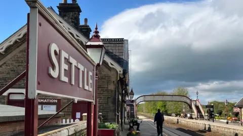 The railway station at Settle with a red sign with white lettering in the foreground. There are two platforms in the shot, with two people waiting on one of them. There is a pedestrian overpass in the background.