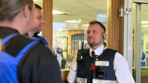 Roger Webb, a white security guard speaking to two other security guards at the entrance of A&E