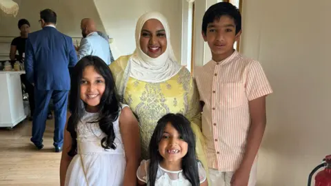 Tania Naseer Tania Naseer, wearing a yellow dress with white hijab, smiling with her son and two daughters