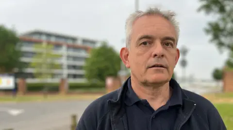 Alan has grey hair and is wearing a black coloured polo shirt and zip up jumper. Behind him, in soft focus, is the Carruthers Court building.