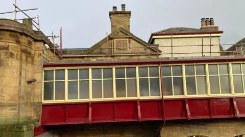 Charles Heslett/BBC The outside of a Yorkshire stone building with a metal walkway painted in maroon and yellow. A date stone on a chimney stack above reads 1885.