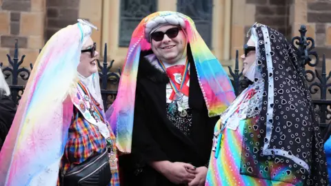 Three mourners dressed as colourful nuns, wearing rainbow habits and sunglasses and rainbow lanyards around their necks