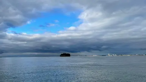 jez A big blue hole in the cloudy skies above a round fort sat in the middle of the Solent. In the background is Portsmouth with the white Spinnaker Tower in Portsmouth highlighted by sunlight.