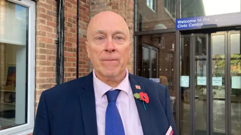 A man stood in front of a council building, wearing a blue suit jacket, pink shirt and blue tie. 