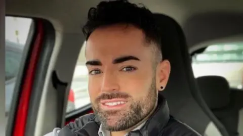 Karl Whitcomb Karl, who has dark hair and a short dark beard, is sitting in a car wearing a dark gilet and a grey shirt.