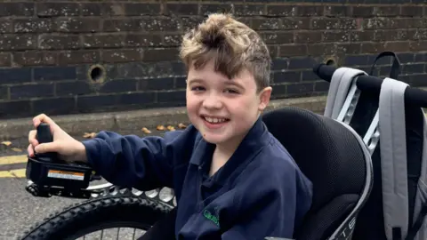 Ben is wearing a blue top and has short brown hair. He is sat in a large electric wheelchair.