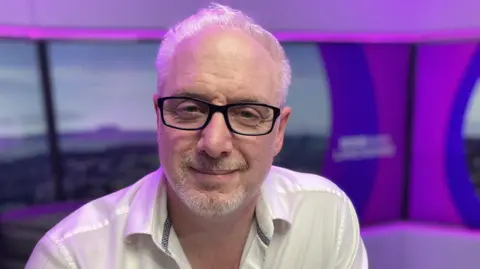 Dr Chris sits in a radio studio wearing a white shirt and black glasses. He has short white hair.