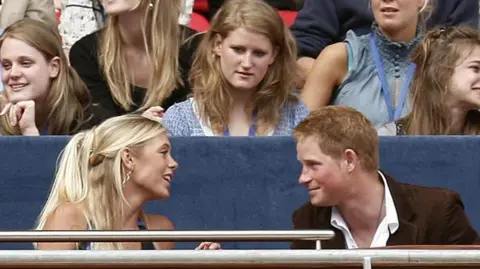 PA Media Prince Harry at the age of 22, wearing a brown suit jacket and white shirt, speaks to then-girlfriend Chelsy Davy, who has blonde hair and wears a sleeveless top, at a concert