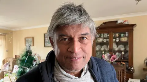 Stephen looking down the camera, he's wearing a navy fleece with a beige jumper underneath. He's stood in a living room, with a Christmas tree just visible over one shoulder. The walls are beige and you can see a cupboard with china on display against one wall.