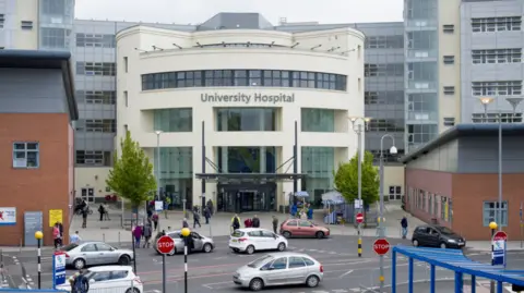A large modern hospital building with the words University Hospital on the front. The main entrance has tall glass windows and a number of people are outside. In front of the hospital is a street with cars. 

