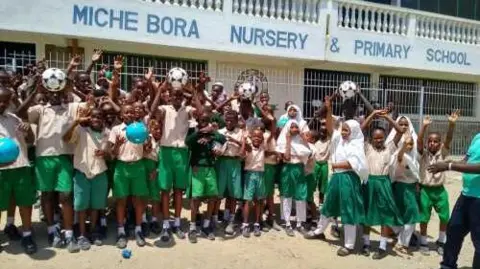 Rita Fowler Children outside the school in Kenya wearing green shorts or skirts and holding footballs above their heads