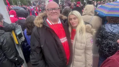 Stephen Brady, who has a bald head and wears black framed glasses and is wearing a black coat over a Liverpool football top, poses with his arm around his daughter Elle-May, who has long blonde hair and is wearing a light beige coat over a Liverpool top. They are standing in a large crowd of fans. 